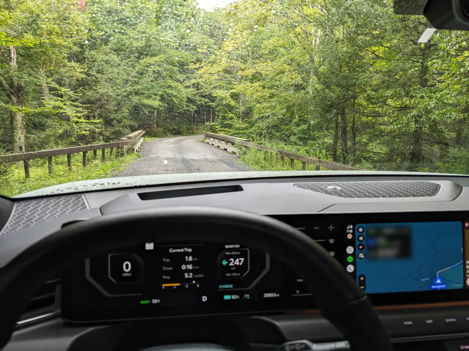 View through the Kia EV9 windshield of a wet gravel forest road leading to a one-lane bridge